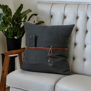 May include: A dark gray throw pillow with a brown leather strap and a pocket for holding a remote control and glasses.