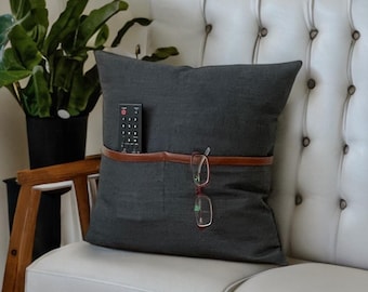 Puede incluir: Una almohada gris oscuro con una correa de cuero marrón y un bolsillo para guardar un mando a distancia y gafas.