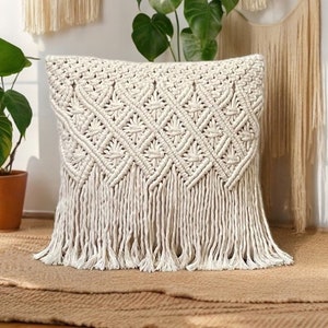 Peut inclure: Un coussin en macramé blanc avec un motif en losange et des franges. Le coussin est posé sur un tapis tissé brun.