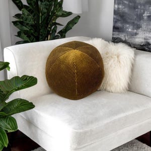 Op de afbeelding: Een ronde, olijfgroene fluwelen kussen met een chevronpatroon ligt op een witte bank. Een pluizig, wit kussen en een schilderij zijn op de achtergrond te zien. Groene planten geven de scène een natuurlijke uitstraling.