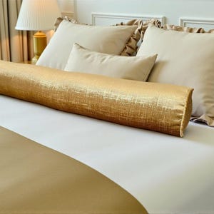 Könnte beinhalten: Ein goldfarbenes Nackenkissen mit strukturierter Oberfläche liegt auf einem Bett mit weißen Laken. Beige Kissen mit Rüschenkanten und ein kleineres Kissen befinden sich dahinter. Die Tagesdecke ist goldfarben.