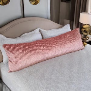 Puede incluir: Una almohada lumbar de terciopelo rosa con un sutil patrón texturizado se encuentra sobre una cama blanca con almohadas blancas. La almohada está colocada sobre una cama con una funda nórdica blanca.