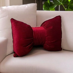 Puede incluir: Una almohada de terciopelo rojo en forma de lazo con ribete blanco. La almohada está en un sofá blanco.