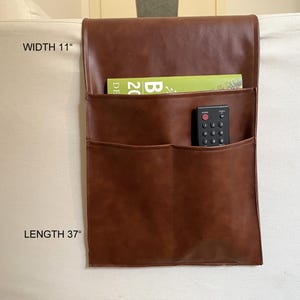May include: Brown leather couch armrest organiser with two pockets and a remote control holder. The organiser measures 37 inches in length and 11 inches in width.