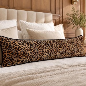 May include: A long, rectangular bolster pillow featuring a prominent leopard print pattern in shades of brown, tan, and black. The pillow has a distinct black piped edge that outlines its elongated shape, resting horizontally across a bed.