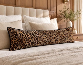 Puede incluir: Un cojín alargado y rectangular con un llamativo estampado de leopardo en tonos marrón, beige y negro, rematado con un borde ribeteado en color negro, colocado horizontalmente sobre la parte delantera de una cama con ropa de cama neutra.