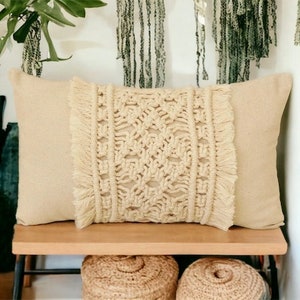 May include: A beige throw pillow with a cream-coloured macrame design and fringe trim. The pillow is sitting on a wooden bench.
