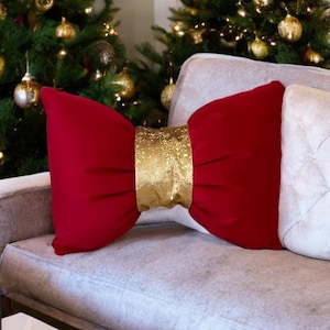 Puede incluir: Un cojín decorativo rojo en forma de lazo con una banda dorada, colocado sobre un sofá gris. Hay una almohada blanca al lado. El fondo incluye un árbol de Navidad con adornos dorados.