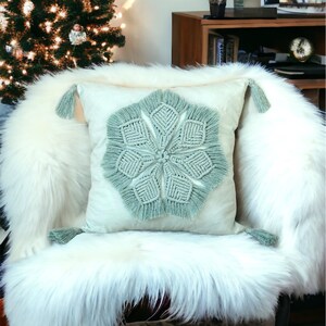Pode incluir: Uma almofada verde claro com um design decorativo de macramé e borlas. A almofada está em uma cadeira de pele sintética branca.