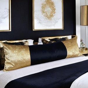 May include: A black and gold lumbar pillow with a velvet texture. The pillow is on a bed with a white and black patterned duvet cover.