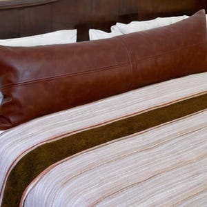 May include: A brown leather body pillow on a white and brown striped bedspread.