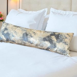May include: A long, rectangular decorative pillow with a floral pattern in shades of gray, beige, and gold rests on a white bed. Two white pillows are behind it. A vase of pink flowers is on the left.