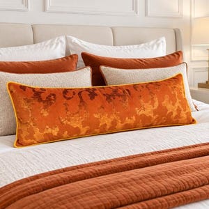 May include: A long rectangular lumbar pillow featuring a burnt orange velvet fabric with a darker orange abstract pattern and yellow piping. It is positioned on a white bedspread alongside other orange and cream-colored decorative pillows.