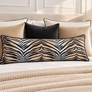 May include: A long rectangular lumbar cushion featuring a black and tan zebra stripe pattern with black piping edges. The cushion is positioned in front of two solid tan square cushions and one solid black square cushion on a white bedspread.