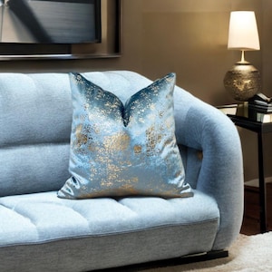 Può includere: Un cuscino quadrato decorativo con un design astratto blu e oro. Il cuscino presenta un effetto sfumato, che passa da un blu più scuro nella parte superiore a un blu più chiaro con accenti dorati. È posto su un divano azzurro.