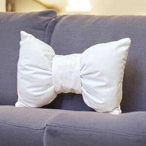 May include: A white bow-shaped pillow sits on a gray couch. The pillow is made of a soft, plush fabric and has a simple, elegant design.
