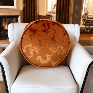 Op de afbeelding: Een ronde, oranje decoratieve kussen met een gouden patroon ligt op een witte fauteuil. Het kussen heeft een textuur en een contrasterende rand. De fauteuil heeft zwarte sierlijsten.