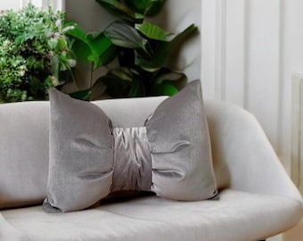 May include: A gray velvet pillow with a large bow in the center. The pillow is sitting on a white couch.
