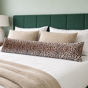 Op de afbeelding: Een bed met een donkergroen hoofdbord, witte lakens en een lang kussen met luipaardprint. Twee beige lendenkussens en twee witte kussens liggen ook op het bed. Een beige deken is over het voeteneinde van het bed gedrapeerd.