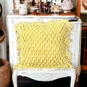 May include: A yellow crocheted throw pillow with fringe trim sits on a white painted chair with ornate legs. The chair is in front of a white dresser with a mirror and decorative items on top.