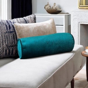 Puede incluir: Un cojín cilíndrico turquesa con textura acanalada descansa sobre un sofá blanco, junto a un cojín rectangular beige y un cojín cuadrado estampado. Los cojines añaden un toque decorativo al salón.