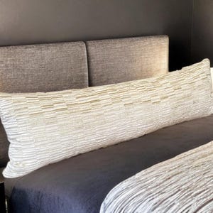 May include: A long, rectangular body pillow with a textured, light-colored cover featuring horizontal stripes. The pillow rests on a bed with a dark blue comforter and a textured, light-colored blanket. A gray headboard is behind the pillow.