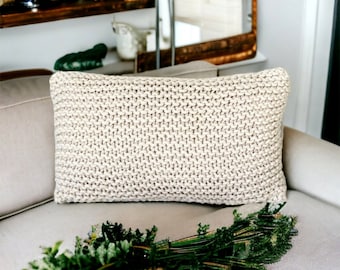 Peut inclure: Un coussin en tricot beige avec un tissage texturé est posé sur un canapé gris clair. Le coussin est net, tandis que le canapé et un bouquet de verdure sont flous à l'arrière-plan.