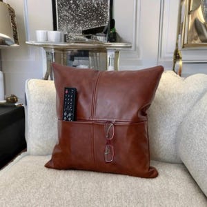 May include: A brown leather pillow with two pockets, one holding a remote control and the other holding a pair of glasses.