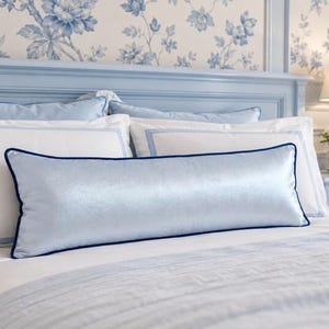 May include: A long, rectangular bolster pillow in a shimmering light blue fabric with a dark blue piped edge. The pillow is positioned horizontally across the front of a bed, resting against two white rectangular pillows with light blue borders.