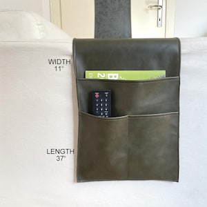 Op de afbeelding: Olijfgroene leren bankarmleuning organizer met drie vakken. De organizer is 94 cm lang en 28 cm breed. Een afstandsbediening en een tijdschrift zijn zichtbaar in de vakken. Ontworpen om over de armleuning te hangen.