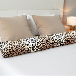 Könnte beinhalten: Ein zylindrisches Nackenkissen mit Leopardenmuster liegt auf einem weißen Bett. Das Kissen hat ein braunes und schwarzes Fleckenmuster. Zwei beige quadratische Kissen und ein weißes Kissen befinden sich dahinter.