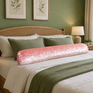 May include: A coral bolster cushion with a floral design lies on a bed with white sheets and a green throw. The bed has a wooden headboard and is in a room with green walls and framed botanical prints.