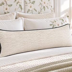 Pode incluir: Uma almofada lombar longa e retangular em tecido creme texturizado. A almofada apresenta um debrum preto distinto ao longo de todas as quatro bordas, criando uma borda limpa e definida contra o material tecido de cor clara da capa.