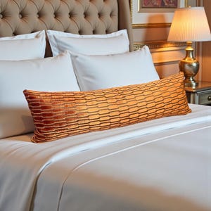 Peut inclure: Un long coussin rectangulaire orange et marron à motif texturé repose sur un lit blanc. Le coussin est long et étroit, avec un motif géométrique répété. Le lit est recouvert de draps blancs et possède une tête de lit capitonnée.