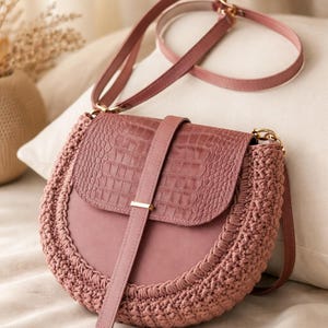 May include: A dusky pink crossbody bag with a crocodile-effect flap and a macrame-style border. The bag has a matching strap and a gold-coloured clasp. The bag is on a cream-coloured surface.