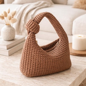 May include: A brown, crocheted handbag with a knotted handle. The bag has a curved shape and a zipper closure. The bag is sitting on a marble table. The background includes a couch, books, and a candle.