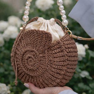 Op de afbeelding: Een bruine, gehaakte handtas met een spiraalvormig ontwerp, met een crèmekleurig interieur met trekkoord. De tas heeft een parelbehandelde handgreep en goudkleurige hardware. De tas wordt door een persoon vastgehouden.