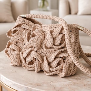 Puede incluir: Bolso beige tejido a mano con ganchillo, con un diseño único y con volantes. El bolso tiene un asa trenzada y un cierre plateado. El bolso está hecho de un material suave y texturizado, y está sobre una mesa de mármol.
