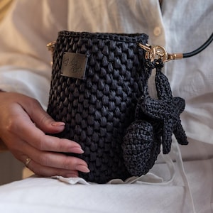 Puede incluir: Bolso bandolera negro de ganchillo con una correa negra y herrajes dorados. El bolso presenta un adorno decorativo de estrella de mar y concha. El bolso se lleva al hombro.