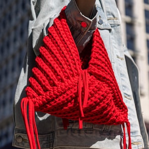 May include: A bright red knitted bag with a distinctive, textured pattern. The bag has long, dangling red cords and is worn over the shoulder. The background features a modern building and a clear blue sky.