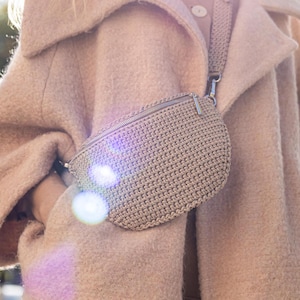 Pode incluir: Uma pochete de crochê bege com um zíper prateado e uma alça longa. A bolsa é usada sobre um casaco bege.
