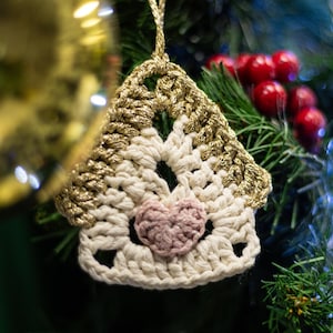 Könnte beinhalten: Ein gehäkelter Haus-Anhänger mit einem rosa Herz in der Mitte, der an einem Weihnachtsbaum mit roten Beeren und goldener Schnur hängt.