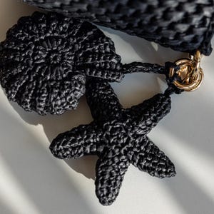 May include: Close-up of a black woven handbag with a matching starfish and seashell charm. The bag features a textured weave and a gold-colored clasp. The charms are attached with a black cord.