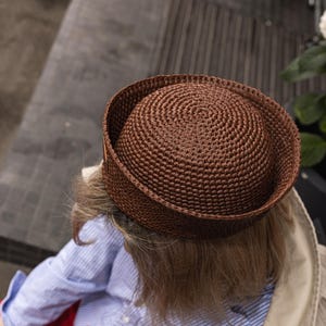 May include: A brown crocheted hat with a rounded crown. The hat is worn by a person wearing a blue and white striped shirt and red trousers. The person is holding a cup of coffee with a foamy top.
