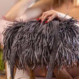 Könnte beinhalten: Eine dunkelgraue Handtasche mit einem dichten, geschichteten Fransen-Design aus dünnen, flachen Streifen. Die Tasche verfügt über einen dicken, geflochtenen Schulterriemen und besteht aus einem strukturierten, gewebten Material.