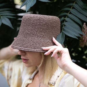 May include: A brown bucket hat crafted from a dense, textured crochet knit material. The hat features a flat top, a structured cylindrical crown, and a short, slightly flared brim that provides a consistent, uniform appearance throughout.