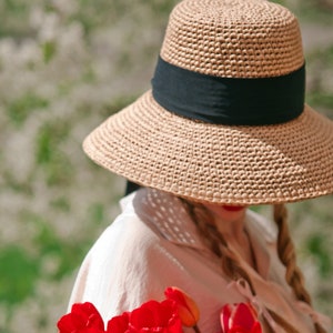 Könnte beinhalten: Eine Frau mit einem breitkrempigen Strohhut mit schwarzem Band hält einen Strauß roter Tulpen.