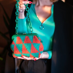Op de afbeelding: Een gehaakte handtas in groen en rood met een geometrisch patroon. De tas heeft een groen frame en een groene ketting. De tas wordt vastgehouden door een persoon die een turquoise top draagt.