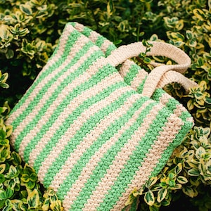 May include: A green and beige striped tote bag with matching handles rests among green and yellow foliage. The bag is made of a woven material, and the stripes run diagonally across the bag.