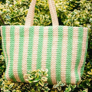 May include: A crocheted tote bag with alternating vertical stripes of light green and beige. The bag has matching beige handles and is set against a backdrop of green and yellow foliage.